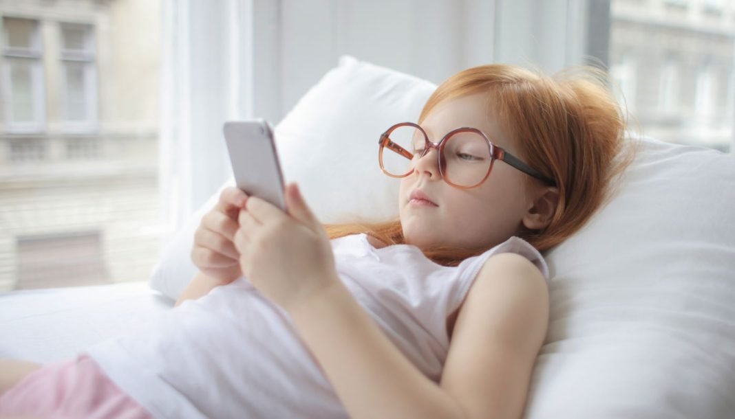 En pige med rødt hår og store briller ligger på en pude og kigger på en smartphone.