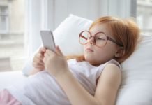 En pige med rødt hår og store briller ligger på en pude og kigger på en smartphone.