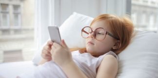 En pige med rødt hår og store briller ligger på en pude og kigger på en smartphone.