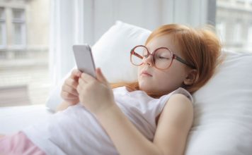 Sådan sælger du din gamle mobil i Danmark En pige med rødt hår og store briller ligger på en pude og kigger på en smartphone.